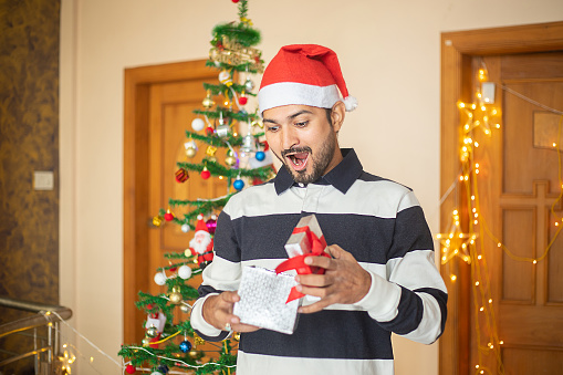 Surprised amazed excited young man holding opened present box wearing santa hat at home, celebrating christmas, holiday. decorated tree and lights in the background