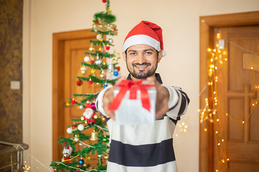 Happy Young Indian man showing gift box in hand celebrating christmas, new year, holiday, party, winter, december. quarantine due to covid-19 pandemic.