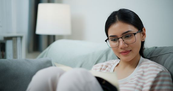 Footage of young Asian woman in glasses sitting on comfortable sofa and enjoy to reading book in living room alone. single woman lifestyle