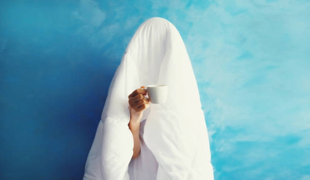 istock Cheerful lazy woman waking up after sleeping covered head with white soft blanket holds cup of coffee on blue background at home 1690263145
