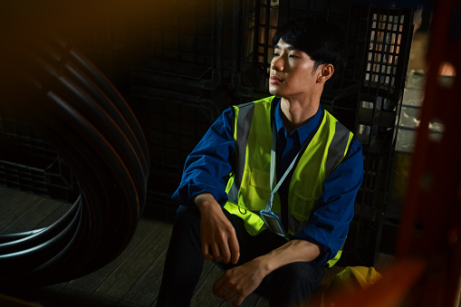 Tired asian male worker sitting on floor in warehouse feeling tried after overwork, burned out or being fired.