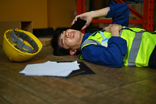 Male worker lying on floor in warehouse after an accident and making an emergency call, asking for first aid team.