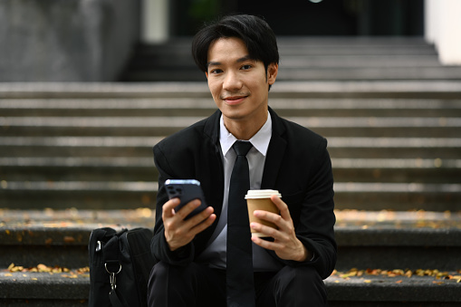 Positive businessman holding disposable cup and using mobile phone outside office building.