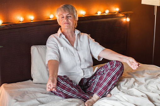 Pretty grey-haired mature woman meditating at home sitting on bed. Yoga, spiritual practice