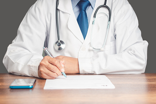 A doctor is holding a pen and writing on a medical prescription while sitting in the hospital. Medical personnel, medicine, and science concept. Quality medical services