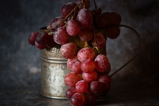 A closeup of a silver can filled with a bunch of ripe red grapes situated on a smooth wooden table.