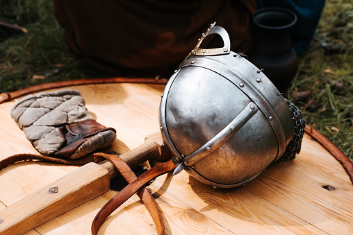 Close-up of a medieval steel helmet lying on an ancient round wooden shield on green grass, outdoors.