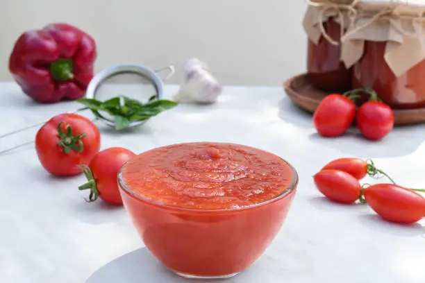 Tomato sauce in a glass bowl. Red tomatoes, sweet pepper, garlic and basil on background. Side view.