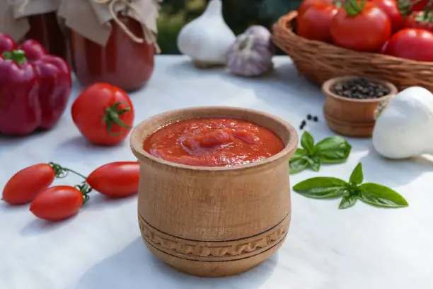 Wooden bowl of tomato sauce with fresh ingredients on white marble background. Side view, place for text copy space.
