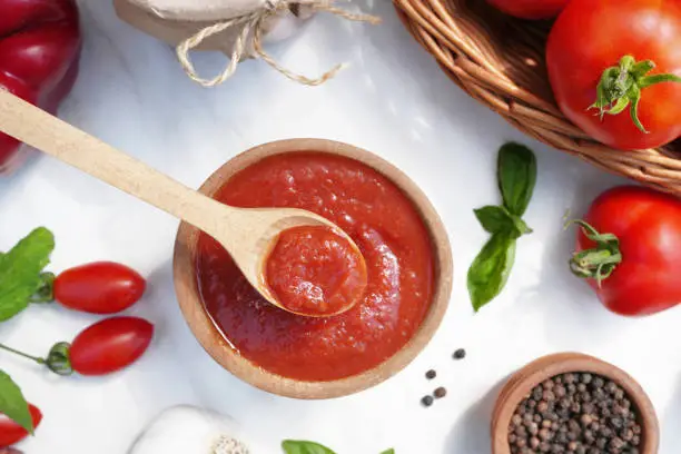 Flat lay composition with tomato sauce in wood spoon and fresh ingredients on white marble background. Top view