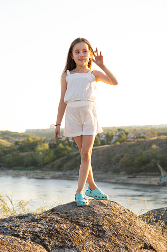 A little five-year-old girl in nature stands on a stone against the backdrop of a river and sky at sunset