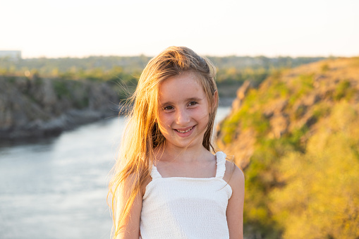 Cute little girl with long hair against the backdrop of a river in nature, smiling, looking away.