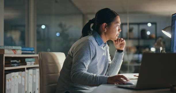 Thinking, overtime and Asian woman at computer in office with research, reading email and tech startup. Internet, analysis and girl typing online report, late at night work and ideas for web design.