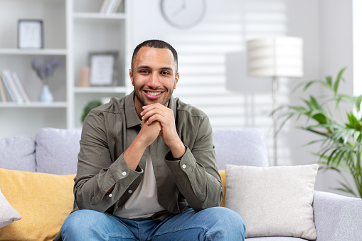 Portrait of a young handsome African-American man sitting on the sofa at home with his hands folded together looking at the camera with a smile.