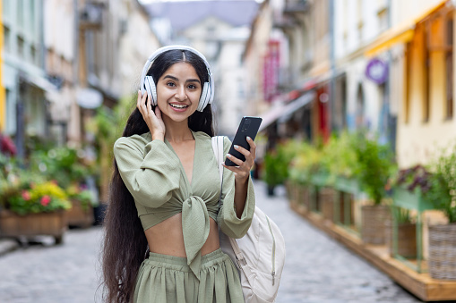 Portrait of a young Indian woman standing on a city street and listening to music in headphones. He holds the phone in his hands and looks at the camera with a smile.