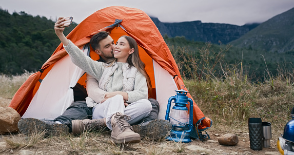Camping, selfie and couple in hug, kiss and relax in nature with love, happy and memory. Campsite, tent and man embrace woman travel influencer for profile picture, social media or podcast photo