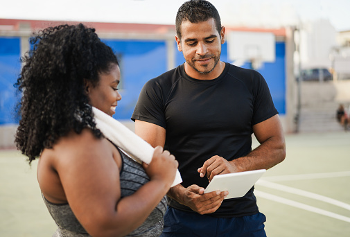 Curvy woman talking with her personal trainer while checking tech clipboard outdoor - Sport and plus size people concept - Focus on man coach face