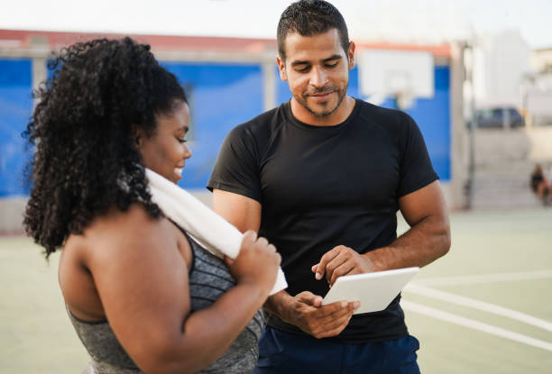 Curvy woman talking with her personal trainer while checking tech clipboard outdoor - Sport and plus size people concept - Focus on man coach face stock photo