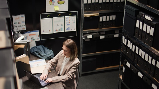 Aerial-view of detective arranging files and evidence, preparing to leave the office. After completing research on criminal case investigation, female investigator closes her laptop on workstation.
