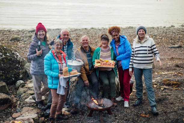 Happy Tourists A group of friends on a staycation together in Torridon, Scotland. They are hanging out in a rural area next to Loch Torridon and are using a fire pit to cook food. They are looking at the camera and smiling while one woman holds a chopping board with freshly langoustine on it and another woman holds a chopping board with sliced bread on it. beach fire scotland stock pictures, royalty-free photos & images