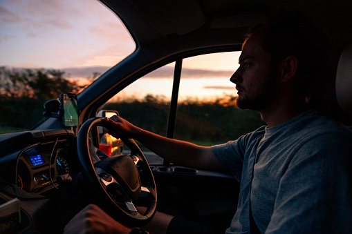 Taken from inside a car, a young man driving a car in Torridon, Scotland during dusk time while the sun is setting. He is looking and concentrating on the road in front of him while holding onto the steering wheel.