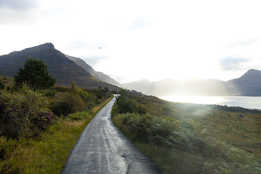 Taken from inside a vehicle, the landscape in Torridon, Scotland on a sunny day. The grassy landscape and mountains can be seen in the distance with a road running towards it. The sunny weather is obscuring the view slightly.