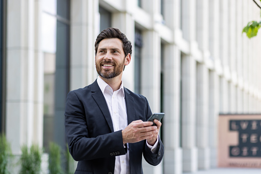 Young cheerful male businessman stands outside office buildings and uses the phone, reads news, types messages, waits for an appointment, looks to the side with a smile.