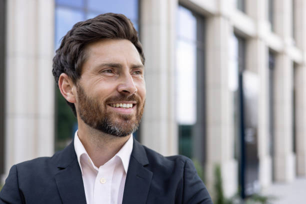 foto ravvicinata di un giovane uomo d'affari bello in un abito da lavoro in piedi fuori da un centro ufficio e distogliendo lo sguardo con un sorriso - close up businessman corporate business side view foto e immagini stock