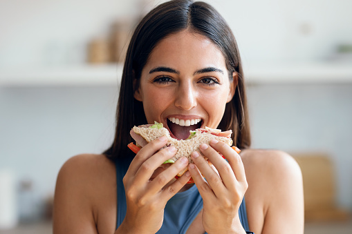 Shot of beautiful sporty woman eating healthy sanwich while looking at camera in the kitchen at home