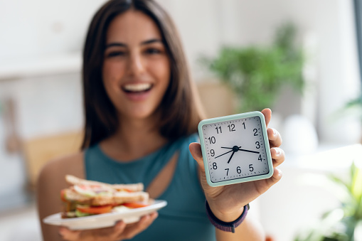 Shot of happy sporty woman waiting for the time to eat while holding clock after fasting in the kitchen at home
