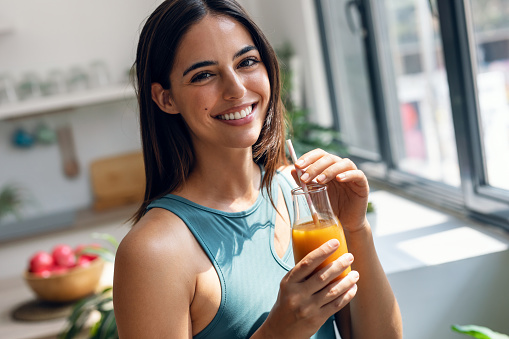 Portrait of beautiful sporty woman drinking healthy orange juice while standing in the kitchen at home