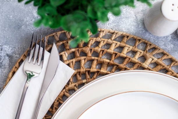 istock Simple dining table setting with white ceramic plate and silver cutlery over rattan round woven placemat on wood background. Close up. 1690602132