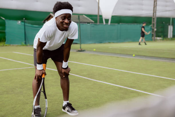 During training on tennis court, young tennis player of African appearance tiredly rested hands on knees. He wants to improve tennis game and practices lot. Man is holding racket and wearing headband