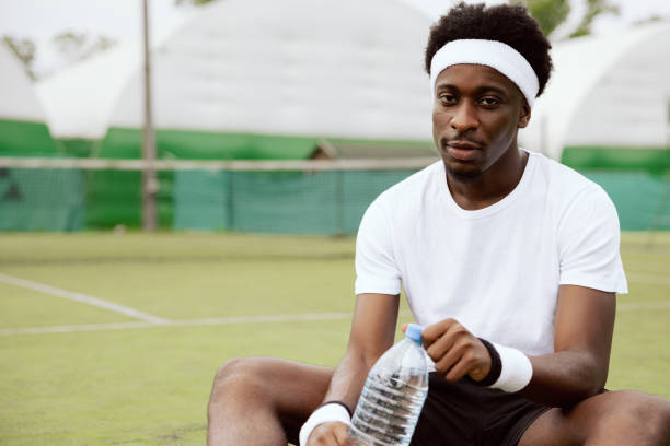 Close-up of the hands of African-looking tennis player sitting on the grass of tennis court and looking tiredly at the camera. After training, man in white t-shirt holds bottle of water and uncorks it