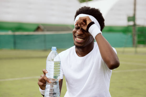Tired, but satisfied with the game, young tennis player took break. He is standing on tennis court and holding bottle of water. Man smiles and wipes the sweat from forehead with hand