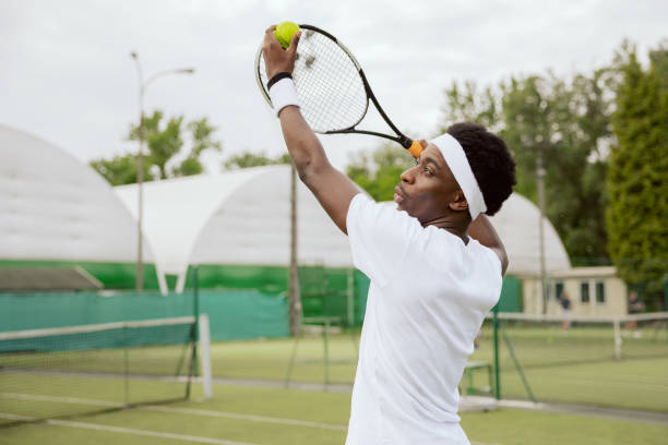 Tennis player is training on the tennis court. Boy holds tennis ball and racket in hands and hits the ball. He is wearing white T-shirt, headband and wristbands. Spotsman makes progress