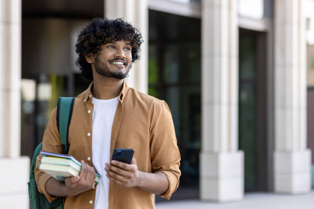 istock Smiling young Indian male student standing outside campus, holding books, backpack and using phone. Looks to the side 1690668638