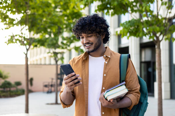istock Young smiling Indian male student came to study, standing outside campus, university and using phone. He was holding books and a backpack 1690668643