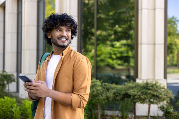 A young Indian man with a backpack and a phone in his hand stands on a city street and looks away with a smile. travels waiting to meet A young Indian man with a backpack and a phone in his hand stands on a city street and looks away with a smile. travels waiting to meet. happy car asian stock pictures, royalty-free photos & images