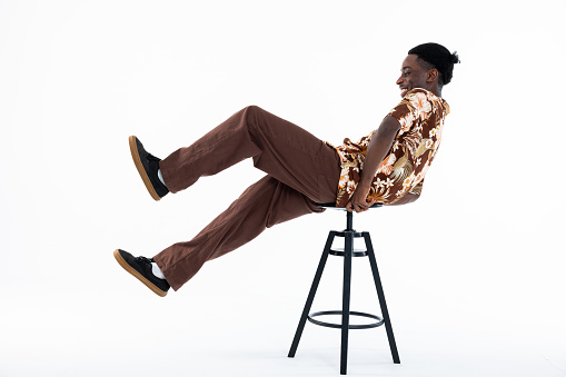 African american man funny posing on chair on white background in studio isolated looking at camera. Wearing colored shirt printed with flowers and brown pants.