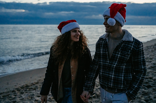 Beautiful young couple walking on the beach at dawn in Nea Flogita, Greece. Wearing Santa hats and celebrating Christmas.