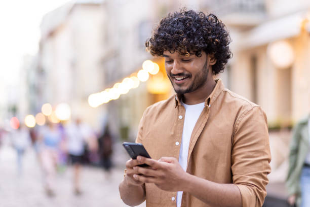Young smiling Indian man standing outside and using phone, texting, chatting, calling