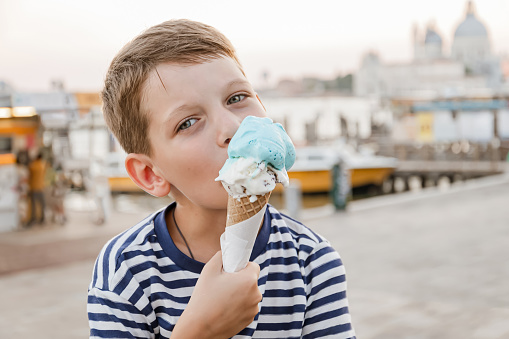 Ð¡ute seven-year-old boy in a striped t-shirt sits on the waterfront in Venice in the evening and eats gelato (ice cream). Tourism and recreation in Europe with children