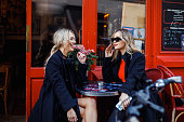 Portrait of two young beautiful women friends wearing black trench coats, sitting at table outside red cafe, talking.