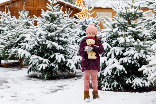Cute little smiling kid girl on christmas tree market. Happy child in winter clothes and toy choosing xmas tree on xmas market with lights on background on winter snow day
