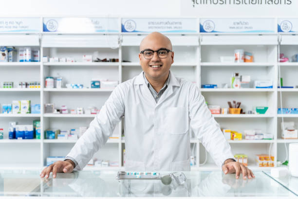 feliz y guapo farmacéutico asiático con anteojos y bata de laboratorio de pie y mirando a la cámara, se siente bien, confiable y orgulloso de su trabajo en la farmacia. - opening bottle pill bottle pill fotografías e imágenes de stock