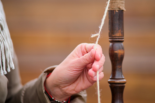 Close up of woman's hands weaving a thread on a spinning wheel. The woman carefully spins the wool into thread using a drop spindle, showcasing her skill in the craft of spinning.