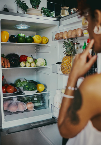 Woman stands in front of an open refrigerator stocked with healthy fruits and vegetables, stock photo