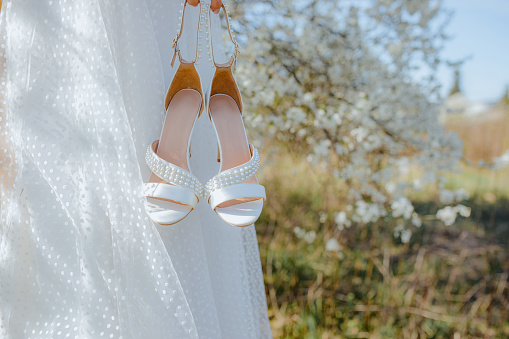 Close up of pair of bridal high heels shoes decorated with small pearls in hand of female in sumptuous wedding gown standing in front of white cherry blossom. Bride holding footwear while walking.