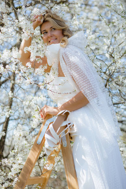 istock Charming lady in wedding attire with big bow on shoulder stand on stepladder among white cherry flowers and smiling. 1690827958